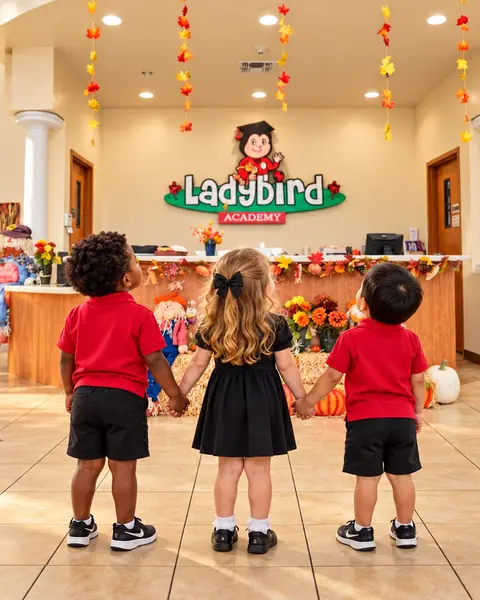 Three preschoolers holding hands in the Ladybird Academy lobby, looking up at the fall-decorated front desk