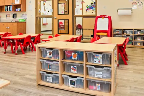 Labeled storage bins of learning materials beside red classroom tables