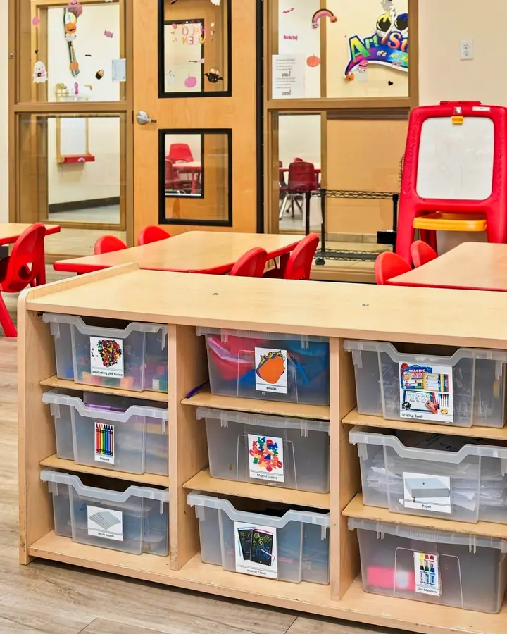 Labeled storage bins of learning materials beside red classroom tables